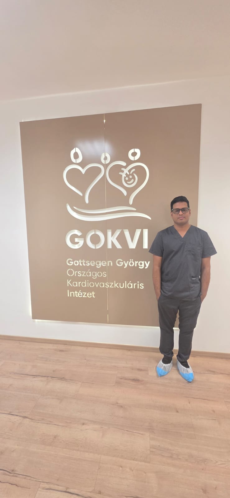 Dr. Gyana Ranjan Nayak in clinical scrubs beside the GOKVI sign during his Structural Cardiac Intervention Fellowship in Budapest.